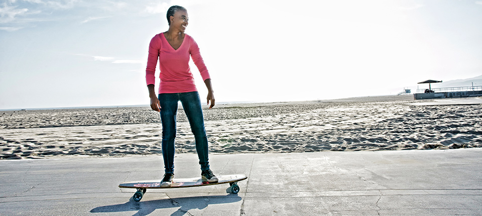 Older Black woman skateboarding by beach